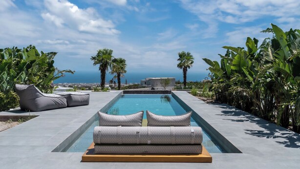 A modern infinity pool with a view of the sea, surrounded by lush greenery and a clear sky. -  Numa Santorini (Santorini)