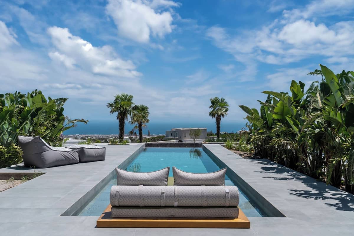 A modern infinity pool with a view of the sea, surrounded by lush greenery and a clear sky.