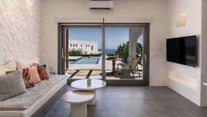 A modern living room with a sofa, two round tables, a TV, and a large sliding glass door leading to an outdoor area with a pool and chairs. -  Numa Santorini (Santorini)