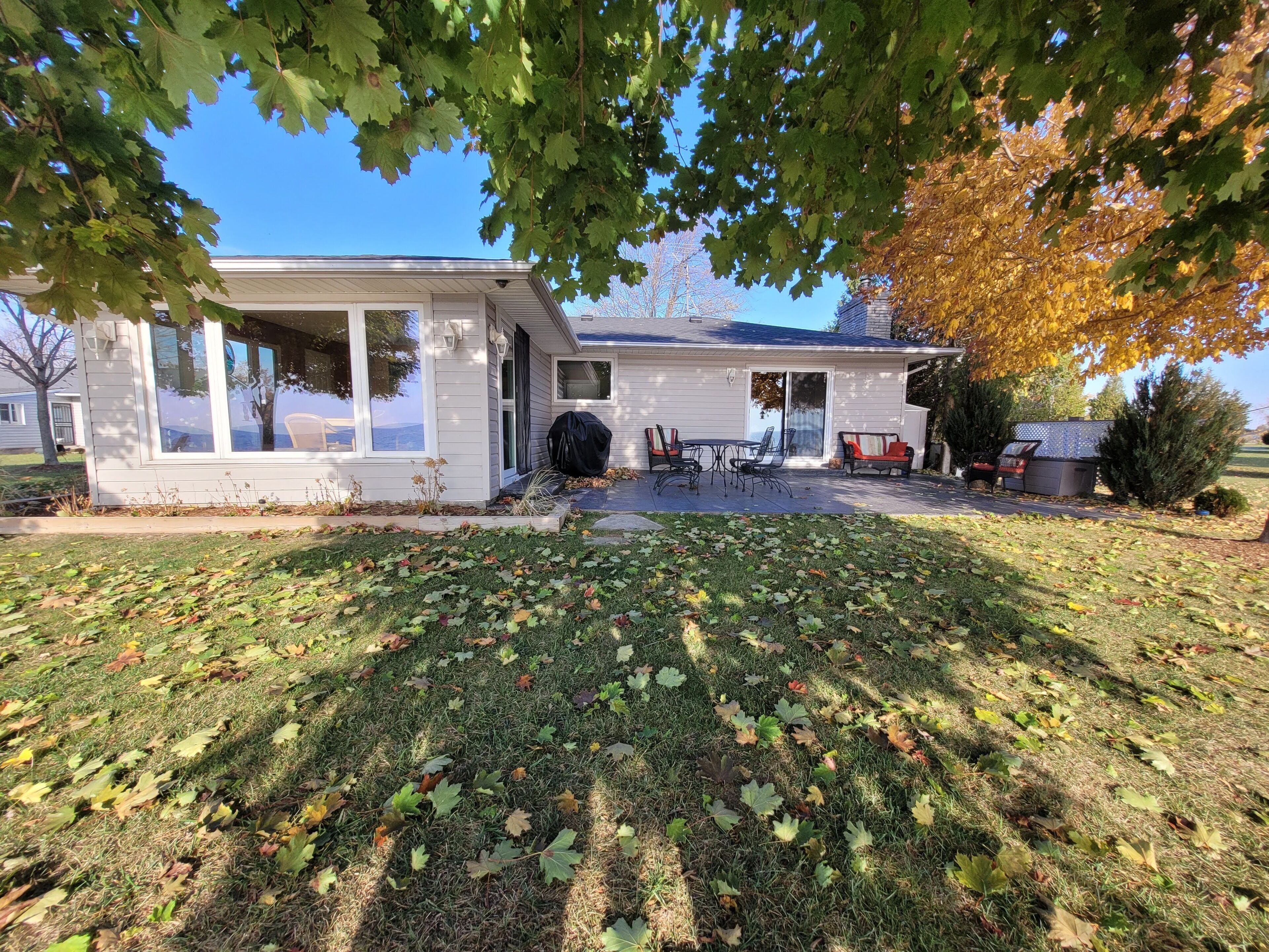 Ein Haus mit überdachter Terrasse, einem Rasen mit verstreuten Blättern und einem Baum mit Herbstlaub.