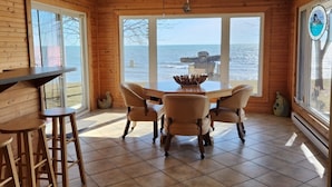 Une pièce aux murs en bois, au sol carrelé, dotée d’un ventilateur de plafond, d’une table avec des chaises et de grandes fenêtres donnant sur la mer.