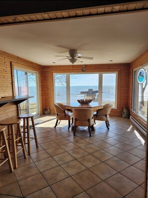 Una habitación con paredes de madera, suelo de baldosas, ventilador de techo, una mesa con sillas y ventanas amplias con vistas al mar.