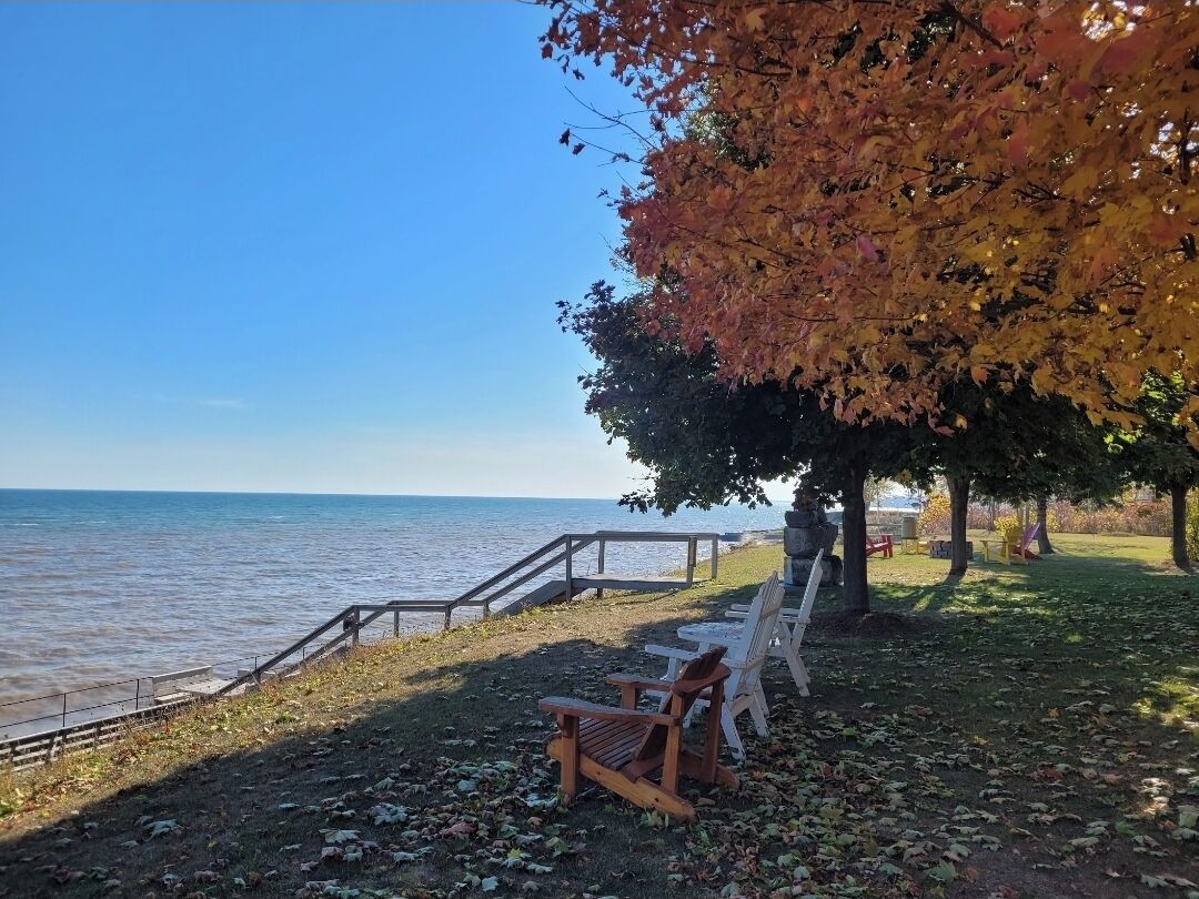 Un parque frente al agua con un banco y sillas de madera, un camino de piedra y vista al agua.