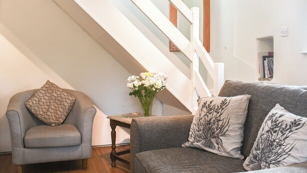 Una sala de estar con un sillón gris, un sofá gris con cojines estampados, una mesa de centro de madera y una escalera con barandilla blanca.