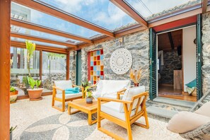 An outdoor seating area with wooden furniture, a stone wall, and a glass door leading to a room.