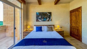 A bedroom with a large bed, two bedside tables with lamps, a wooden door, and a view through a glass door. - Sunset Serenity - Four Bedroom Villa, Sleeps 8 (San Lawrenz)