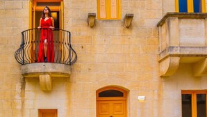 A woman in a red dress standing on a balcony with a wrought iron railing. The building has a stone facade with yellow doors and windows. - Sunset Serenity - Four Bedroom Villa, Sleeps 8 (San Lawrenz)