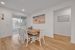 A dining area with a wooden table and white chairs, a painting on the wall, and a window with blinds.