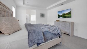 Ein Schlafzimmer mit einem Bett, einem Schminktisch, einem an der Wand montierten Fernseher und einer Tür zu einem anderen Raum.
