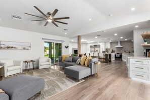 A spacious living area with a grey sofa, a white armchair, and a grey ottoman. There is a kitchen in the background with white cabinets and a stainless steel refrigerator.