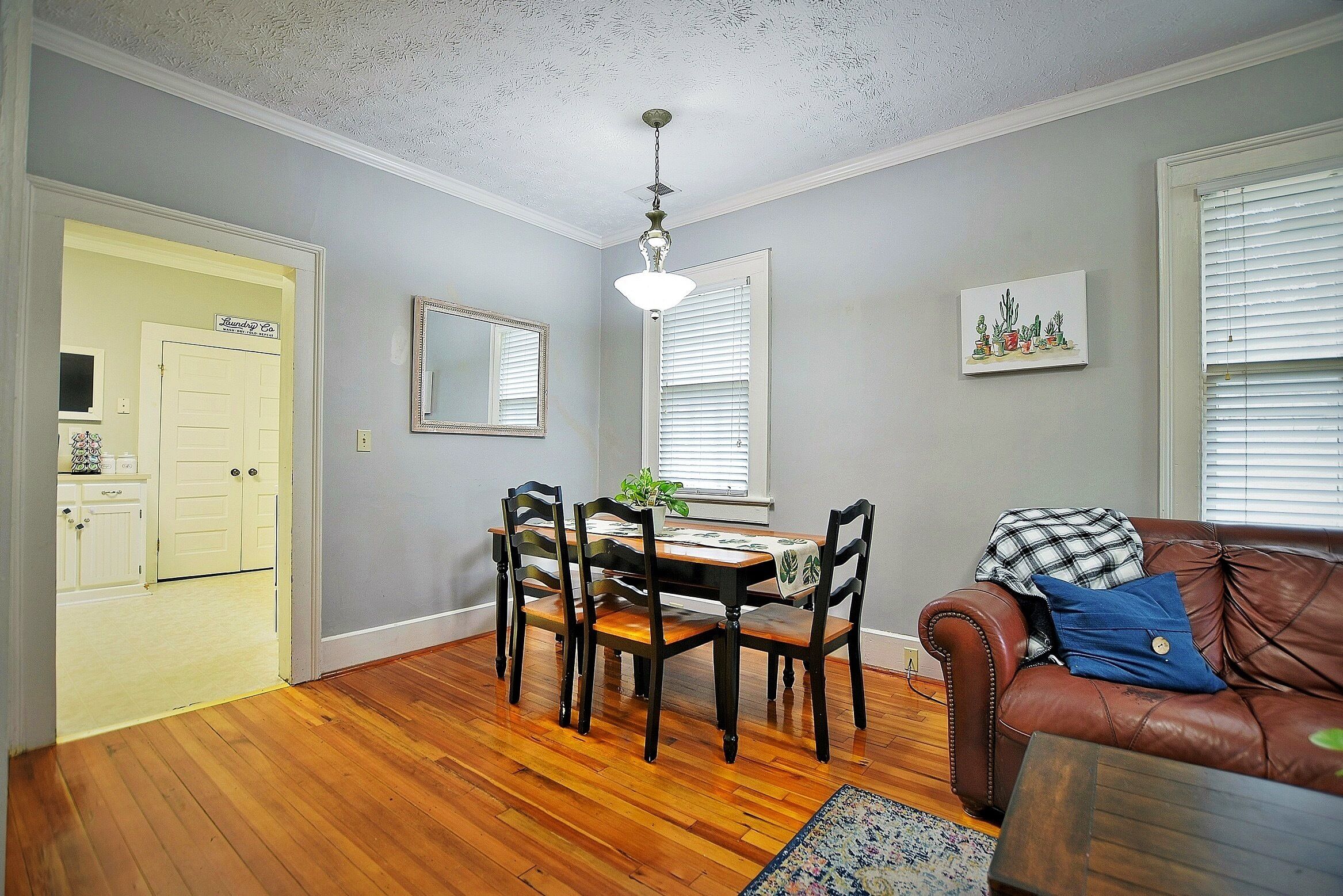 A dining area with a wooden table and chairs, a chandelier, and a brown leather couch with a blue pillow.