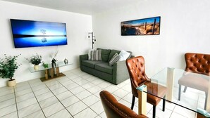 A modern living room with a green sofa, leather armchairs, a glass coffee table, and two wall-mounted TVs displaying nature scenes. - Bayfront  apartment with Breathtaking views.   1 bdr w/ balcony . Free Parking (North Bay Village)