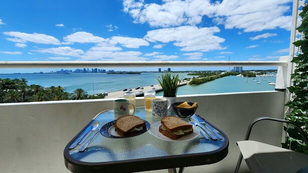 A balcony with a table set for two, overlooking a marina and city skyline. - Bayfront  apartment with Breathtaking views.   1 bdr w/ balcony . Free Parking (North Bay Village)