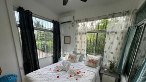 A bedroom with a bed, a nightstand, a blue laundry basket, and a window with curtains. - This house is a 2 bedroom(s), 2 bathrooms, located in Ojochal, Provincia de Puntarenas. (Ojochal)