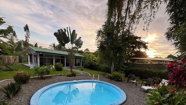 A swimming pool surrounded by gravel, with a house in the background and various plants and trees around. - This house is a 2 bedroom(s), 2 bathrooms, located in Ojochal, Provincia de Puntarenas. (Ojochal)