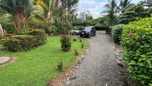 A gravel pathway leading to a vehicle parked in a lush garden with various plants and trees. - This house is a 2 bedroom(s), 2 bathrooms, located in Ojochal, Provincia de Puntarenas. (Ojochal)