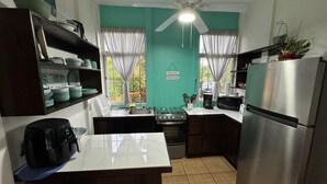 A compact kitchen with a gas stove, microwave, and refrigerator. There are two windows with curtains, a ceiling fan, and a sign on the wall. - This house is a 2 bedroom(s), 2 bathrooms, located in Ojochal, Provincia de Puntarenas. (Ojochal)