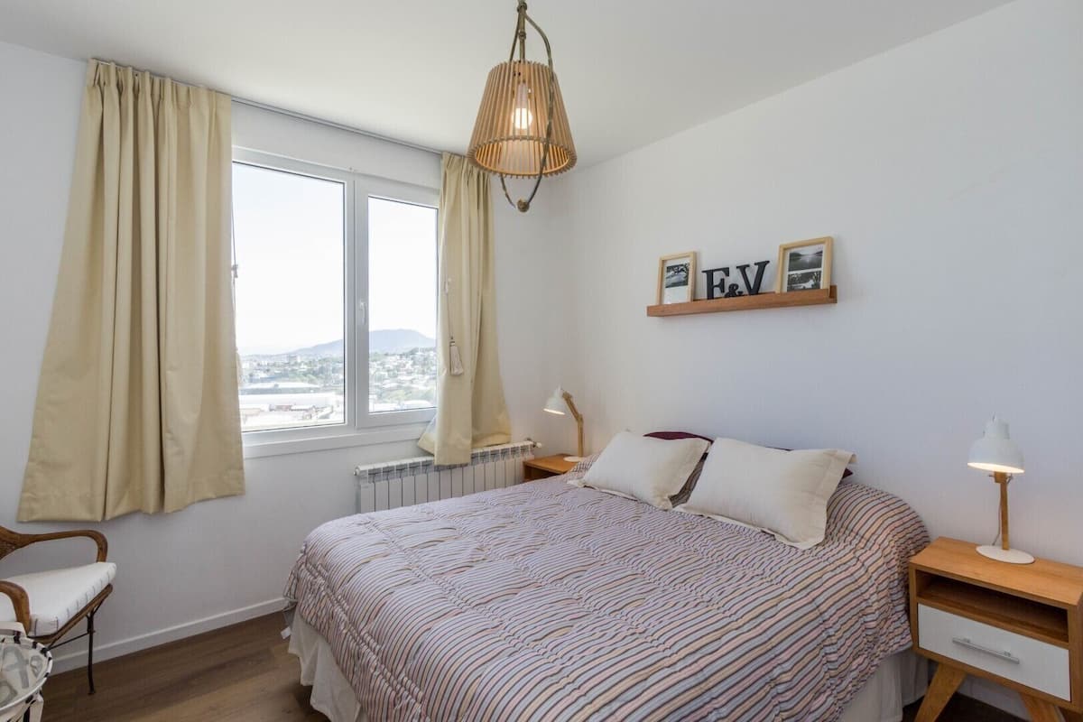 Un dormitorio con una cama, dos mesitas de noche, una silla, una ventana con cortinas, un radiador y una lámpara colgante.