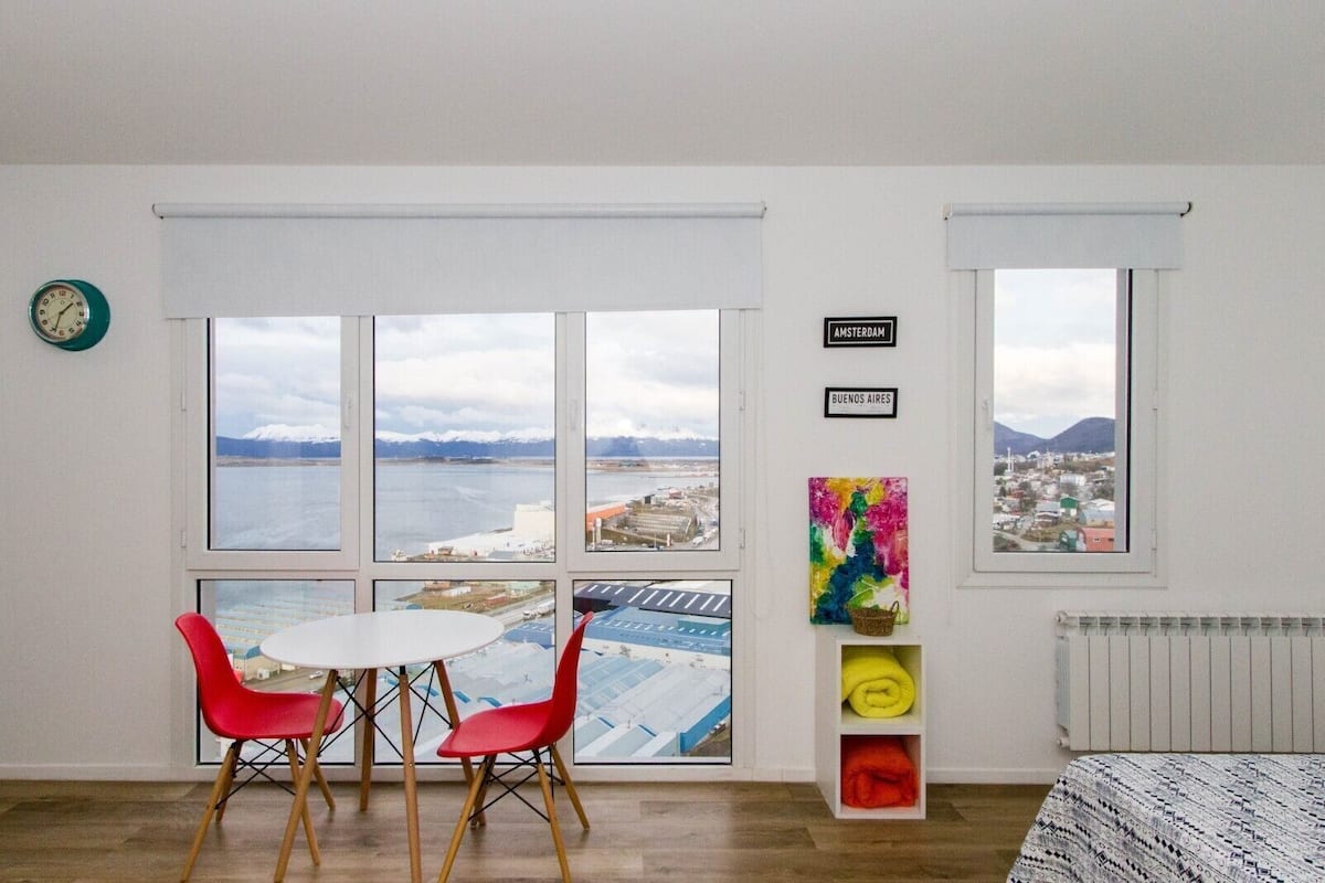 Una habitación con un ventanal grande, una mesa con dos sillas, un radiador y una obra de arte colorida.