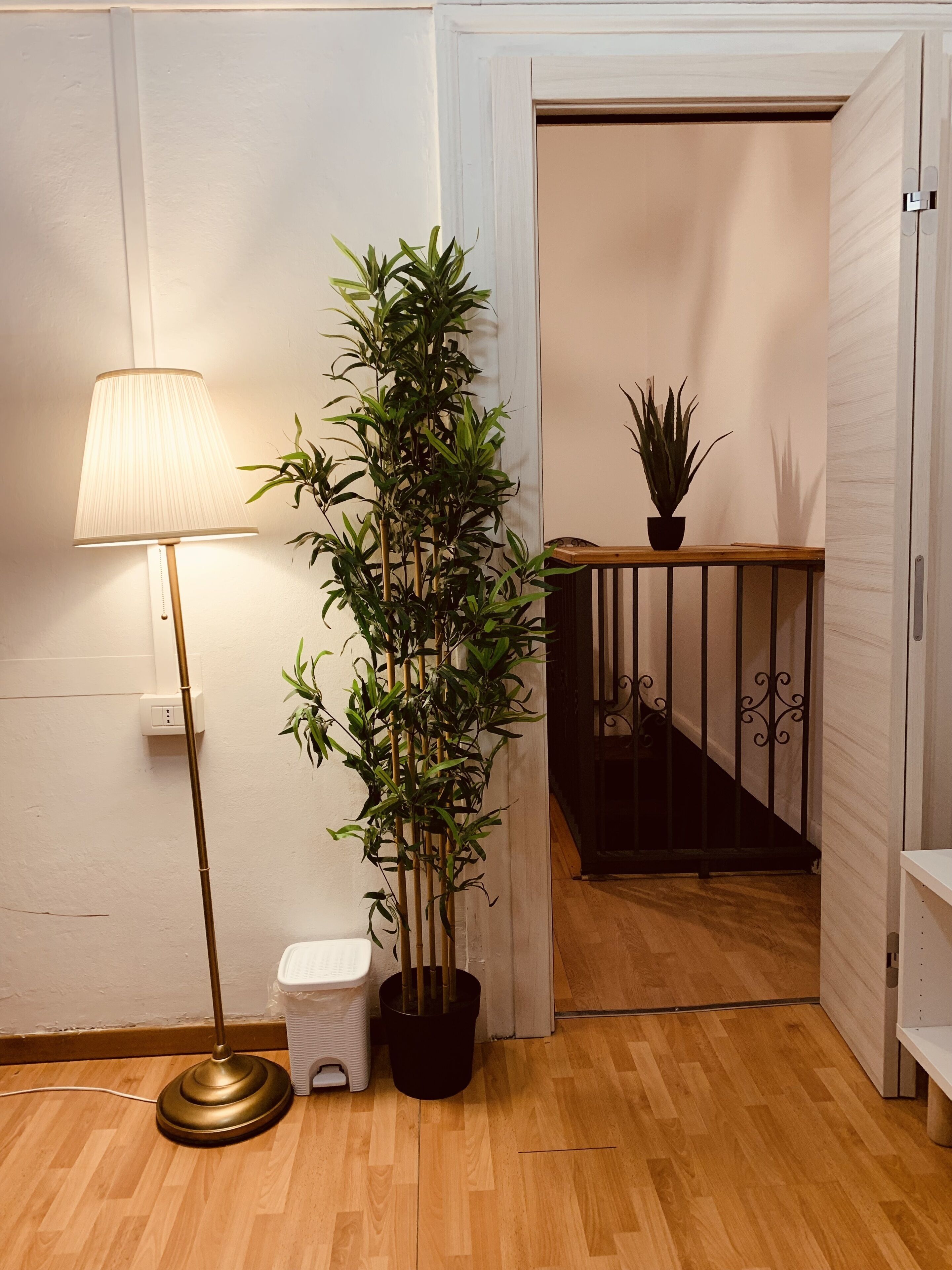 Une pièce avec un lampadaire, une grande plante en pot, un petit tabouret blanc et un escalier menant à une autre pièce.