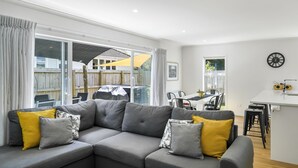 A modern living room with a grey sectional sofa, yellow and grey cushions, a dining area with a white table and chairs, and a kitchen area with a sink and stools. - Contemporary Comfort - Masterton Holiday Home (Masterton)