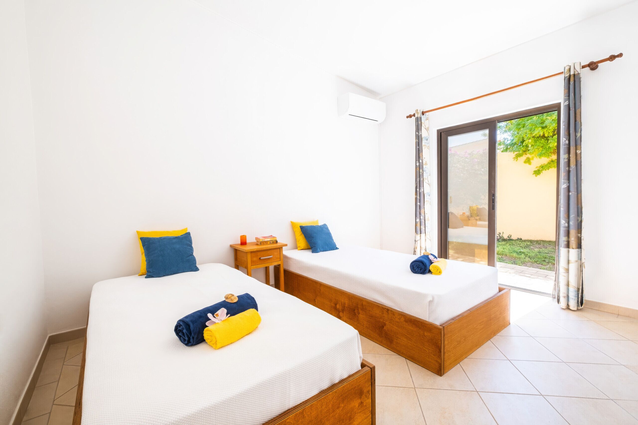 A room with two beds, each with a pillow and a rolled towel. There is a small wooden nightstand with a book and a candle. The room has a sliding glass door leading to an outdoor area.