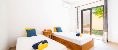 Une chambre avec deux lits, une petite table de chevet en bois et une porte-fenêtre coulissante donnant sur un espace extérieur.