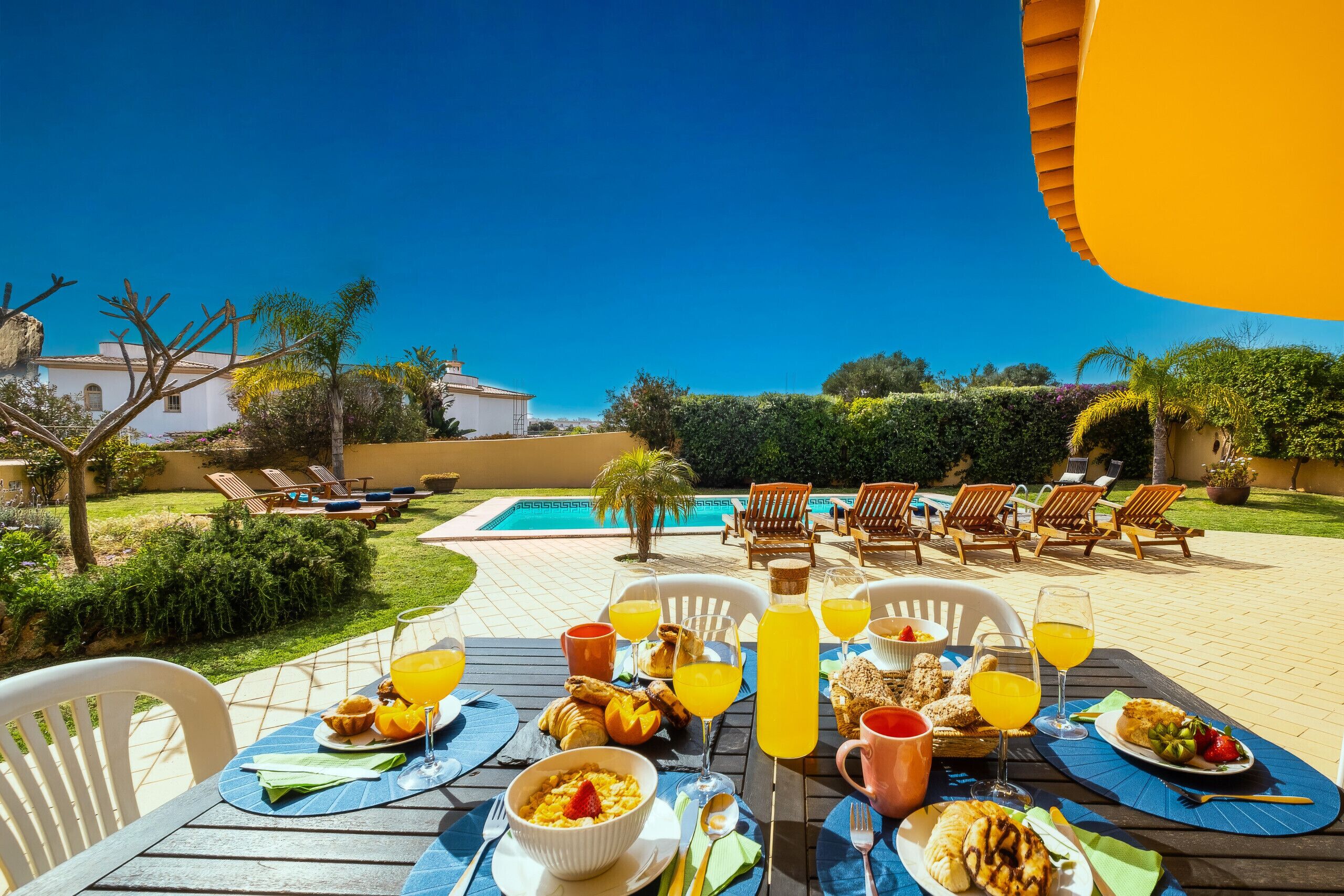 A dining table set outdoors with various breakfast items and drinks, overlooking a swimming pool and garden.