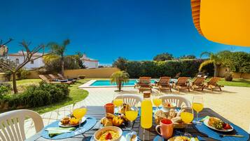 A dining table set outdoors with various breakfast items and drinks, overlooking a swimming pool and garden.