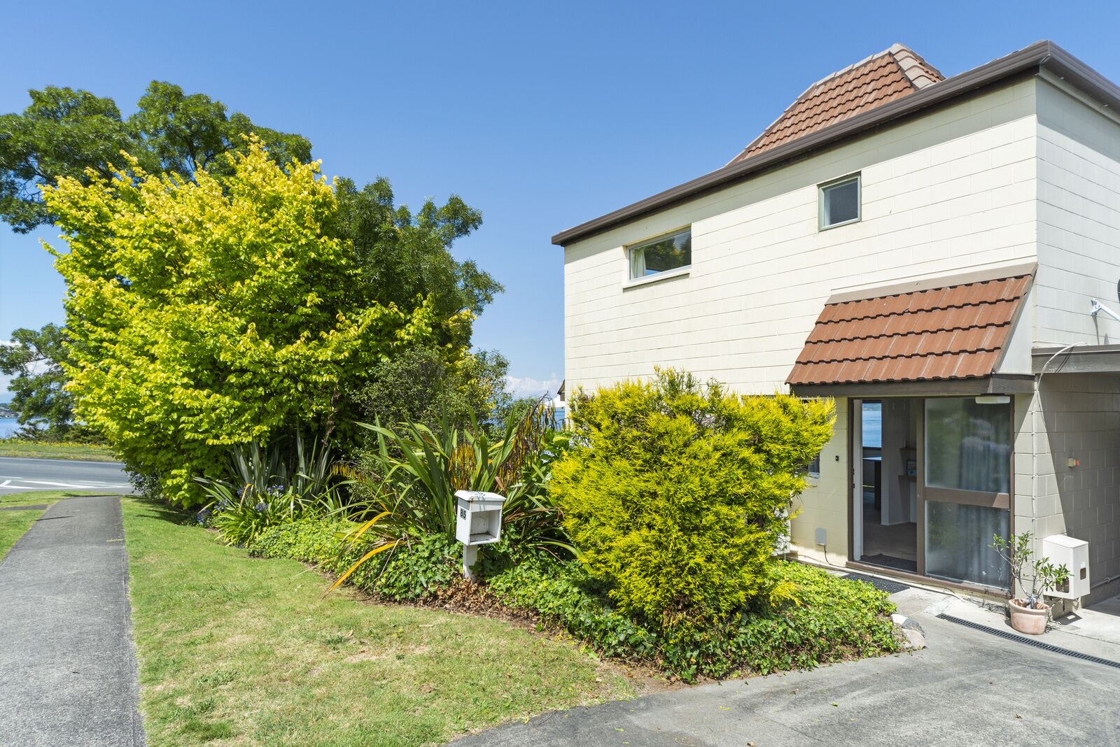 Une maison à deux étages avec un toit recouvert de tuiles rouges, un petit jardin devant avec un arbre et des arbustes, et une boîte aux lettres bien visible.