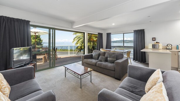 A modern living room with a sofa, coffee table, and a TV. It features large windows with a view of the sea and palm trees. - Lakeside Terrace - Taupo Holiday Home (Taupo)