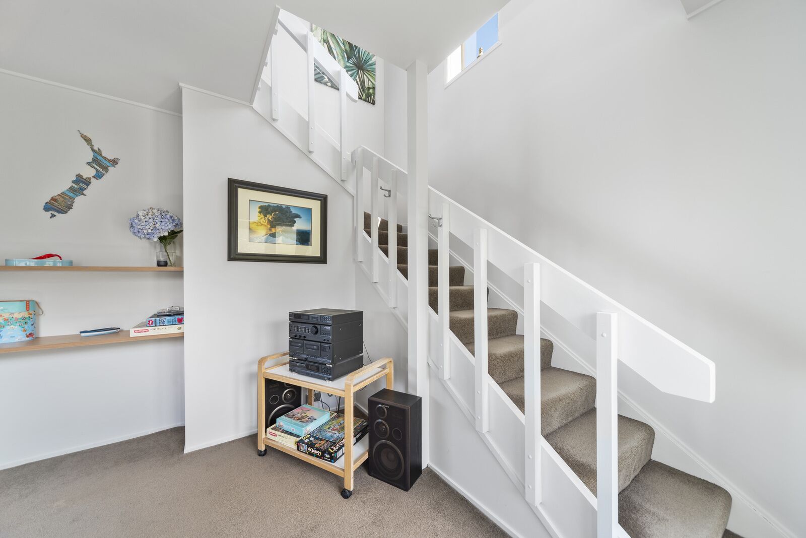 Un escalier avec des rampes blanches et des marches recouvertes d’une moquette beige, une étagère en bois avec des livres et une petite enceinte, et un tableau encadré accroché au mur.