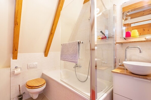 Un baño moderno con inodoro de madera, ducha con cerramiento de vidrio, lavabo blanco y estante de madera con artículos de aseo.