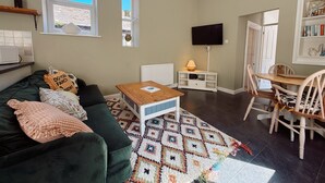 Ein gemütliches Wohnzimmer mit einem grünen Sofa, einem Holztisch, einem gemusterten Teppich, einem Fernseher und einem Bücherregal.