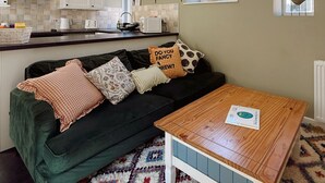 Ein gemütlicher Wohnbereich mit einem dunkelgrünen Sofa, einem gemusterten Teppich und einem Holztisch. Im Hintergrund ist eine Küche mit weißen Schränken und einem Fenster zu sehen.