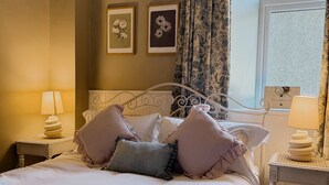 Ein ordentlich bezogenes Bett mit Kissen, ein aufgerolltes Handtuch und ein dekorativer Kopfteil. Zwei Nachttische mit Lampen und gerahmte Blumenbilder an der Wand.