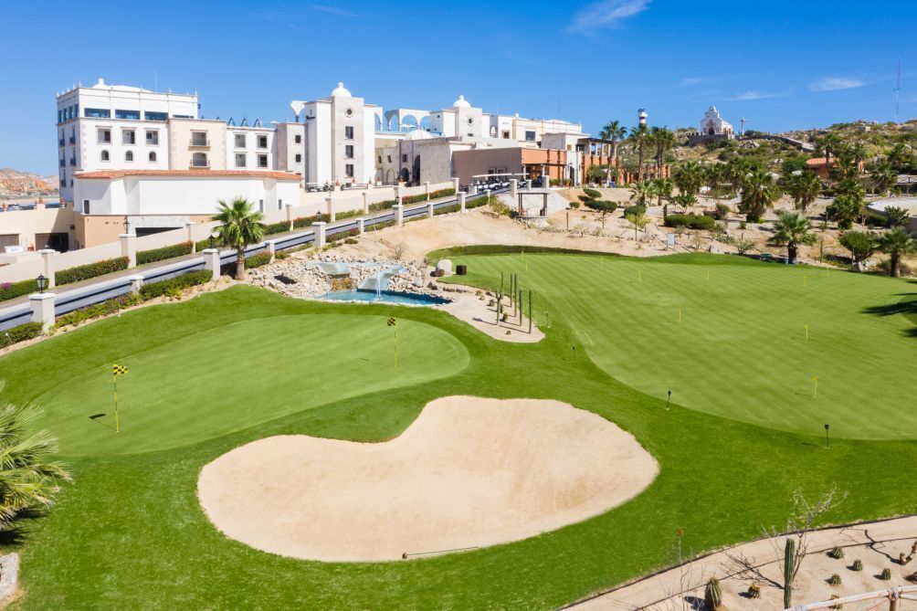 Een golfbaan met een groot zandbunker, goed onderhouden gras en een gebouw met meerdere verdiepingen op de achtergrond.