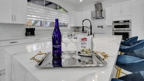A modern kitchen with white cabinets, a large island with a tray of snacks, a bottle of sparkling water, and a bag of wafers. - 4 Bedroom Designer Home Next to Shul and Community Pool (Fort Lauderdale)
