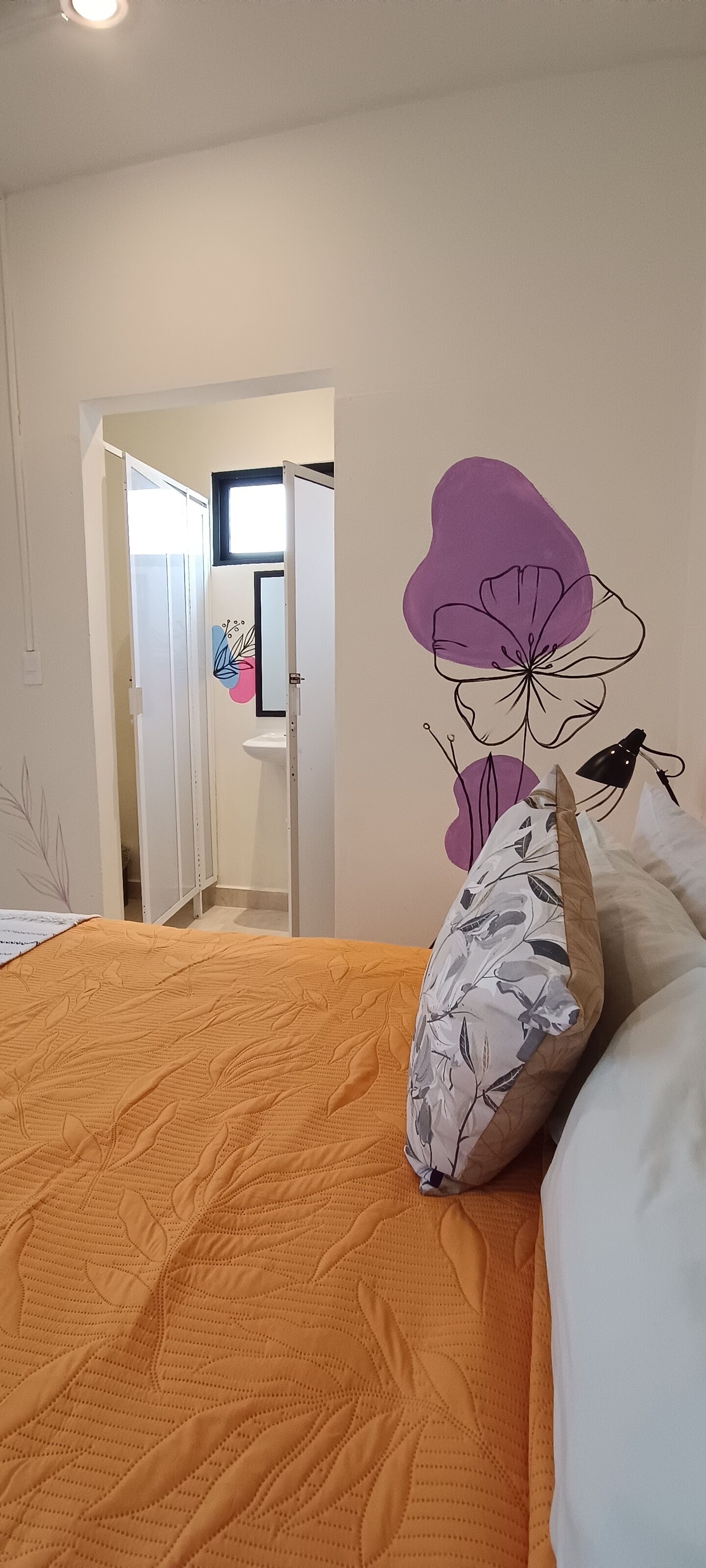 Un dormitorio con una colcha naranja, un cojín floral y un mural en la pared.
