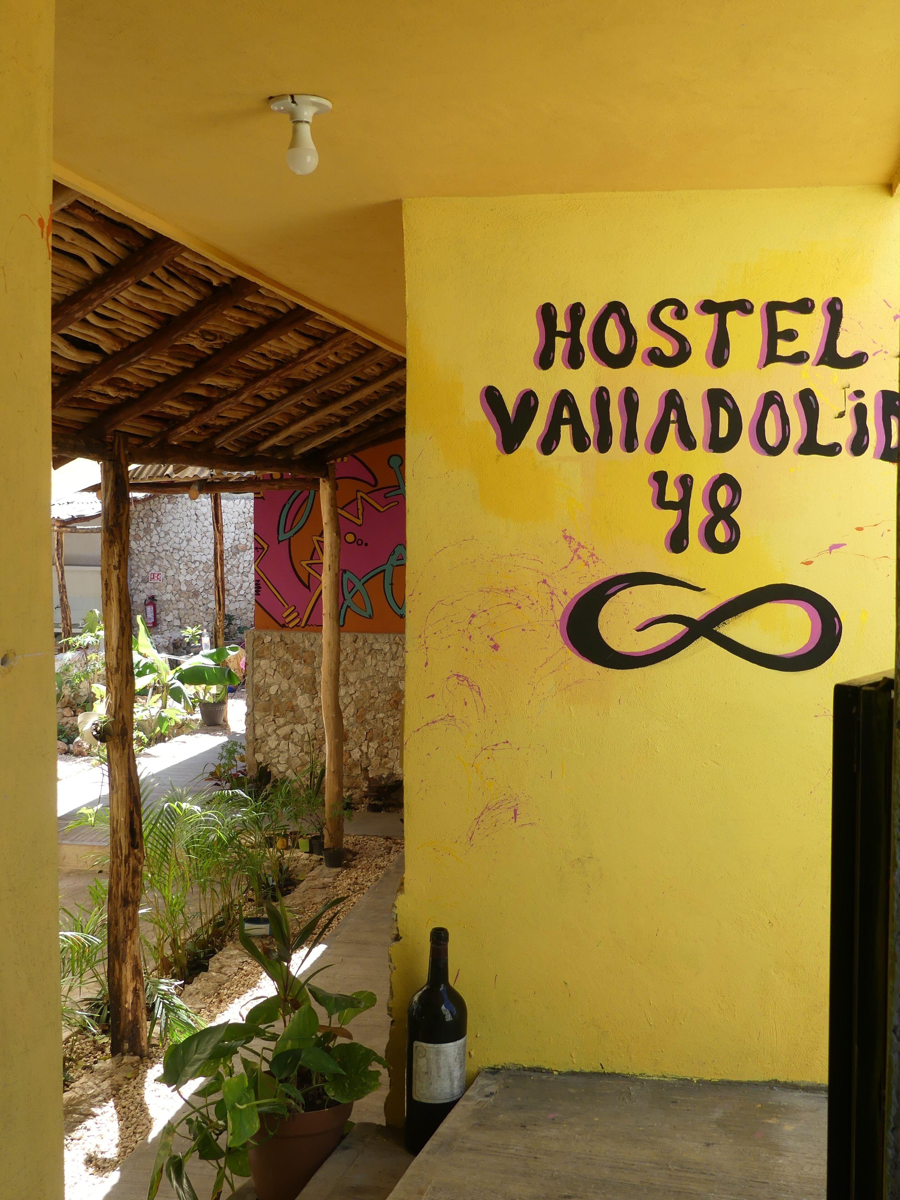 A hostel with a yellow exterior wall, a sign reading "HOSTEL VAIADOLIN 48", and a bottle of wine on the ground.