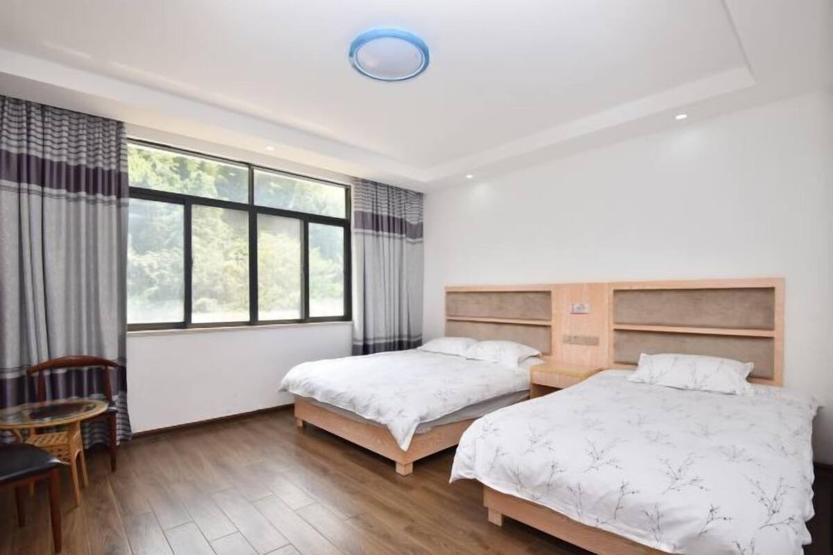 A hotel room with two beds, a wooden floor, a window with curtains, a table with chairs, and a ceiling light.