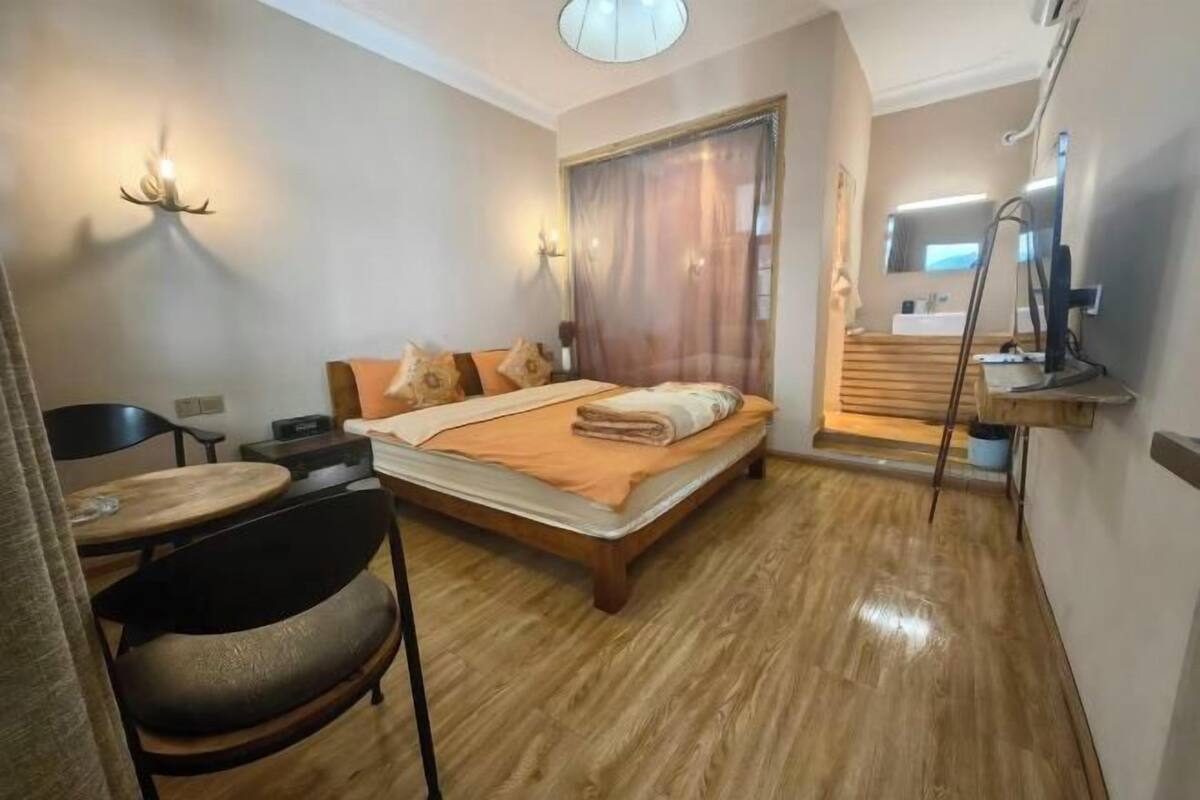 Une chambre à coucher avec un lit, une chaise, une table, un rideau et une salle de bain visible à travers une porte.