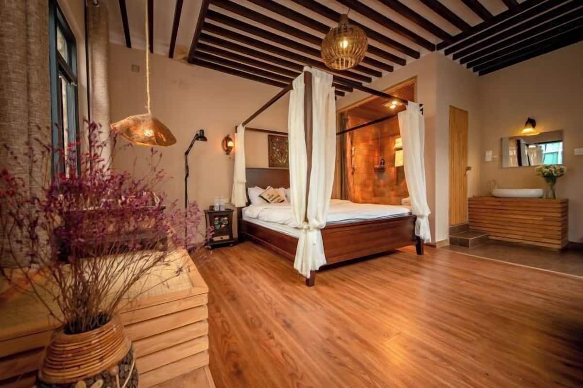 Une chambre à coucher avec un lit à baldaquin, un plancher en bois, une grande fenêtre et une table de chevet avec une lampe.