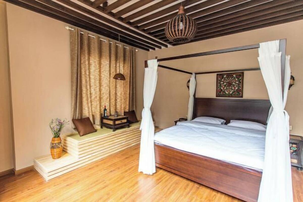 Une chambre avec un plancher en bois, un lit à baldaquin, une table de chevet, une lampe, des rideaux et une petite table sur laquelle est posé un vase.