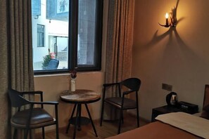 A hotel room with a bed, two chairs, a small table, a window with curtains, and a wall-mounted lamp.