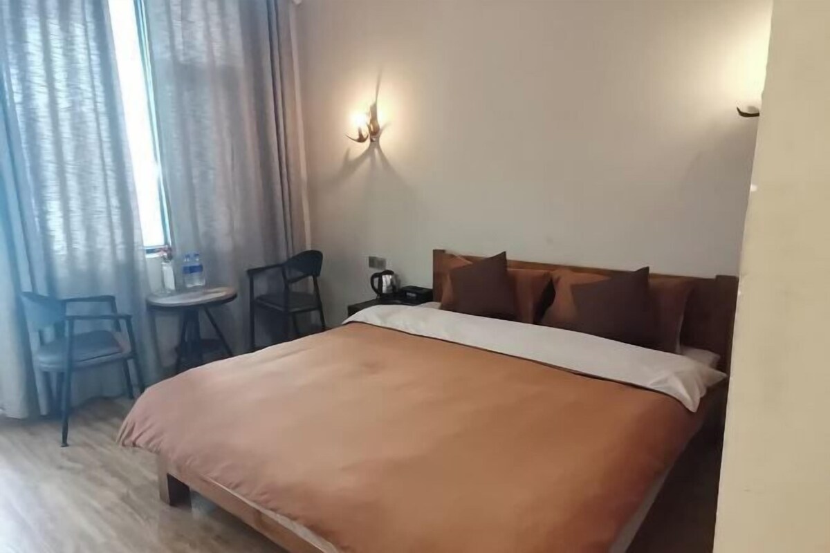 A hotel room with a bed, a small table, a chair, and a wall-mounted lamp.