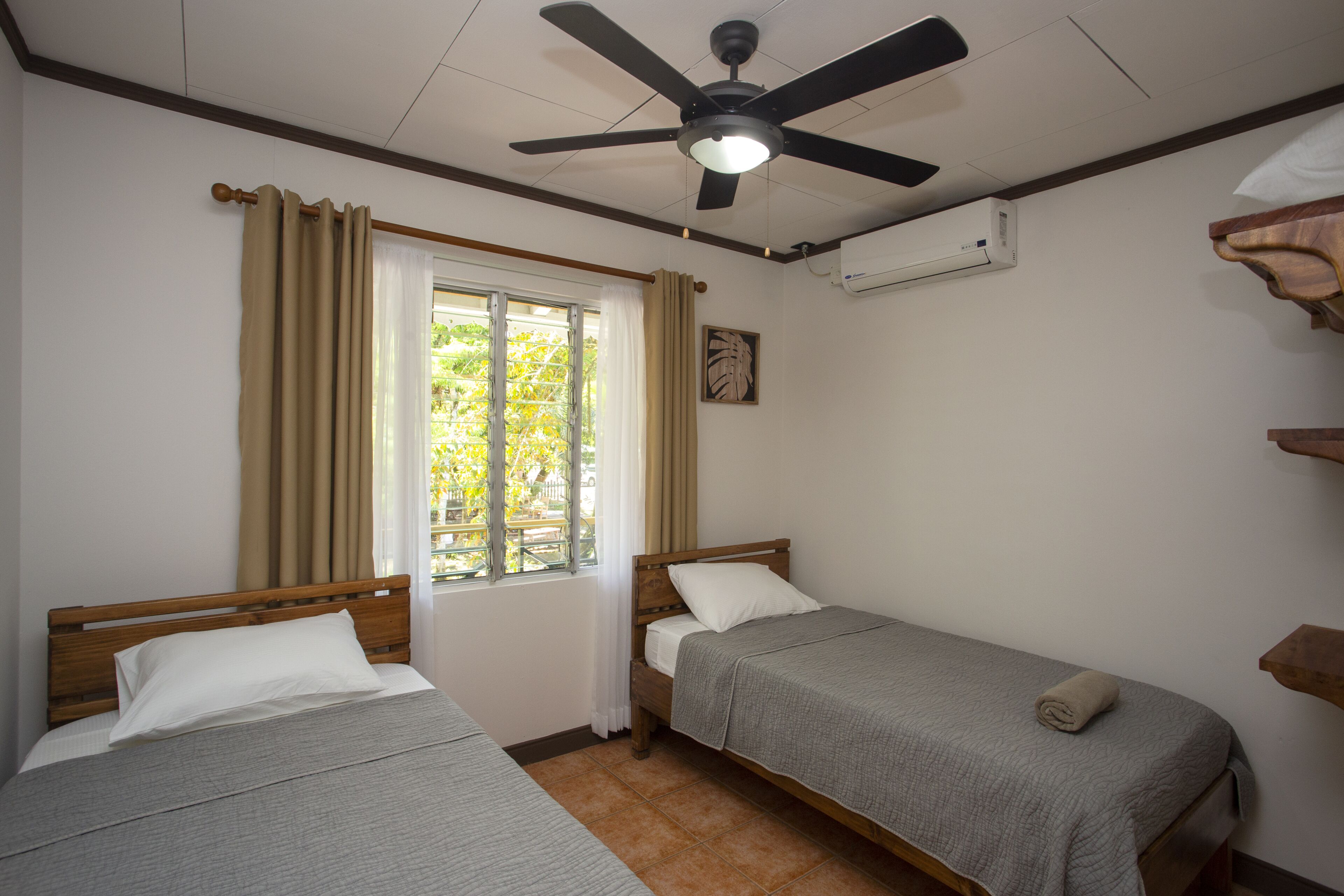 Une chambre avec deux lits simples, un ventilateur de plafond, un appareil de climatisation, une fenêtre avec des rideaux et un petit tableau accroché au mur.