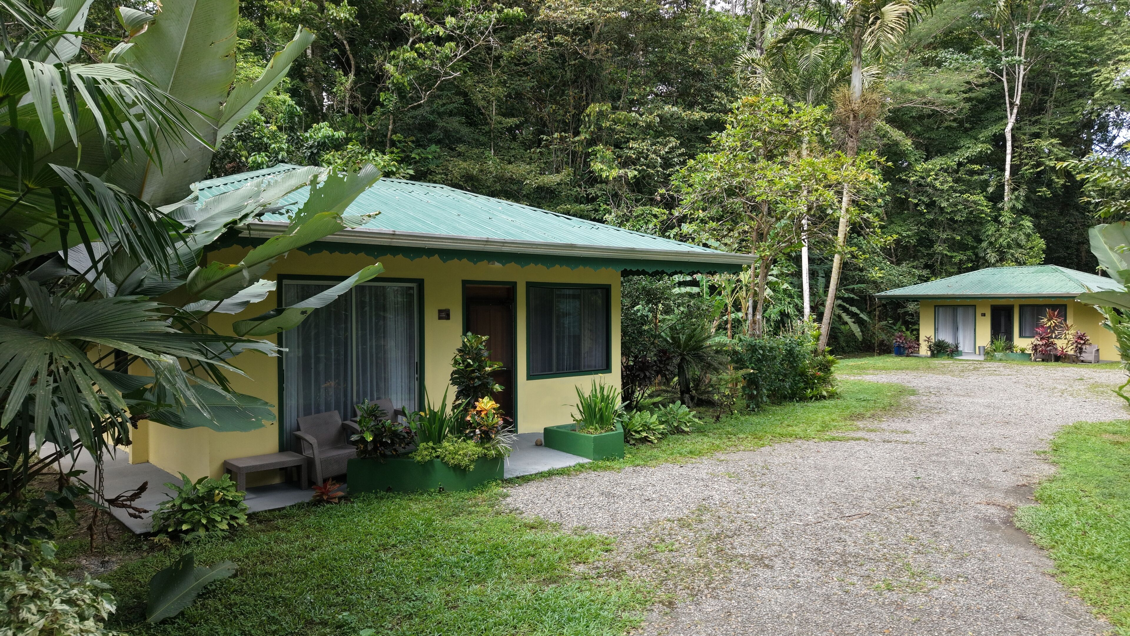 A small, single-story building with a green roof, surrounded by lush greenery and a gravel path.