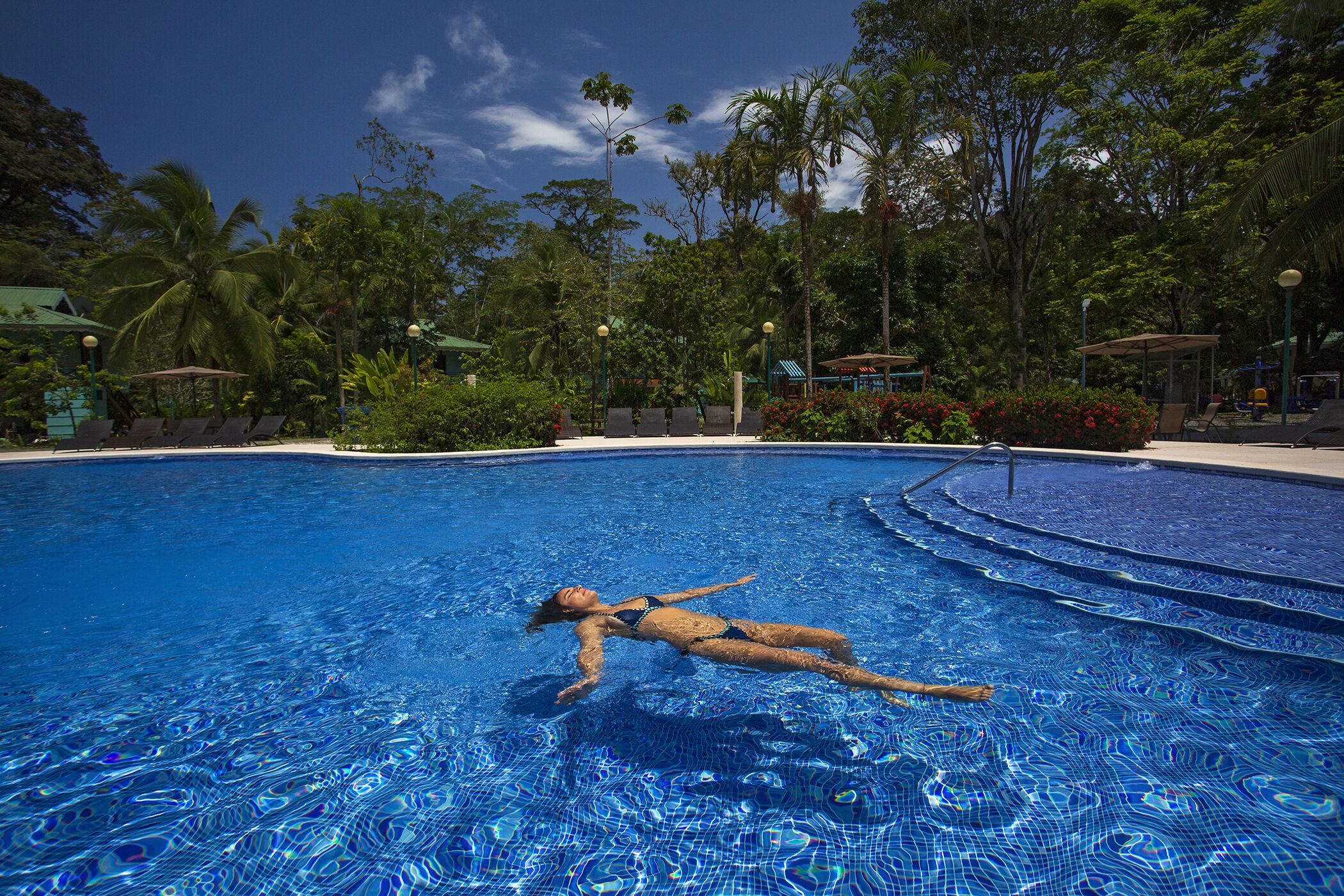 A person floating in a clear blue pool surrounded by lush greenery and palm trees.