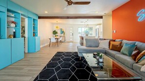 Ein modernes Wohnzimmer mit Sofa, Couchtisch und Deckenventilator. Im Hintergrund ist eine Küchenzeile mit Esstisch und Stühlen zu sehen.